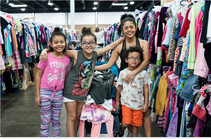 Mom wears baby and holds toddler boy clothes in each hand. Second mom shops behind her from the racks of boys clothes.