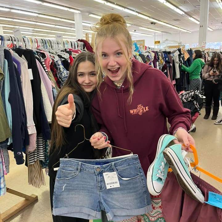 Mom wears baby and holds toddler boy clothes in each hand. Second mom shops behind her from the racks of boys clothes.